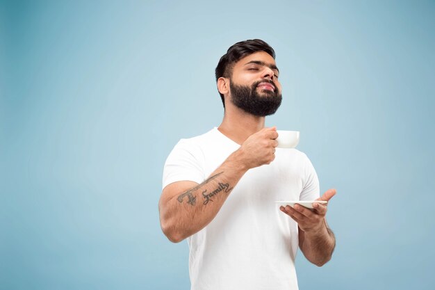 Поясной конец вверх по портрету молодого индусского человека в белой рубашке на синем фоне. Человеческие эмоции, выражение лица, продажи, концепция рекламы. Негативное пространство. С удовольствием пьет кофе или чай.