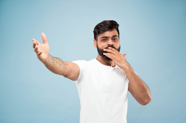 ハーフレングスは、青い背景に白いシャツを着た若いヒンドゥー教の男の肖像画をクローズアップします。人間の感情、顔の表情、販売、広告のコンセプト。ネガティブスペース。幸せで驚いていることを指摘します。