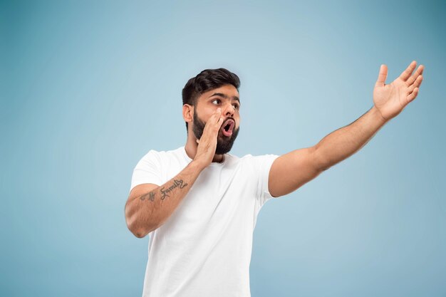 Half-length close up portrait of young hindoo man in white shirt on blue wall. Human emotions, facial expression, ad concept. Negative space. Pointing up being shocked and astonished.