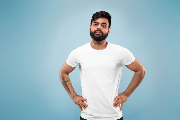 青い壁に白いシャツを着た若いヒンドゥー教の男のハーフレングスのクローズアップの肖像画。人間の感情、顔の表情、広告のコンセプト。ネガティブスペース。ポーズをとって、立って、笑って、落ち着いて見えます。