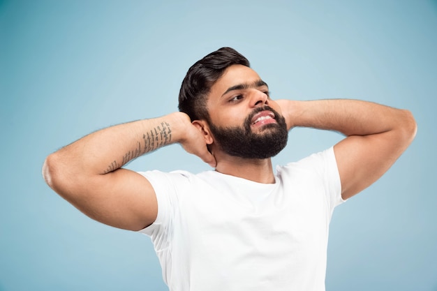 Half-length close up portrait of young hindoo man in white shirt on blue wall. Human emotions, facial expression, ad concept. Negative space. Resting, relaxing, looking calm.