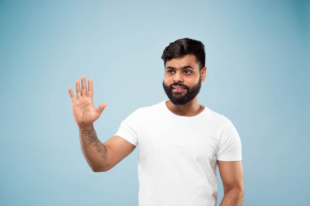 Half-length close up portrait of young hindoo man in white shirt on blue wall. Human emotions, facial expression, ad concept. Negative space. Showing empty space bar, pointing, greeting.