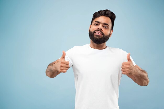 Half-length close up portrait of young hindoo man in white shirt on blue wall. Human emotions, facial expression, ad concept. Negative space. Showing the sign of OK, nice, great. Smiling.