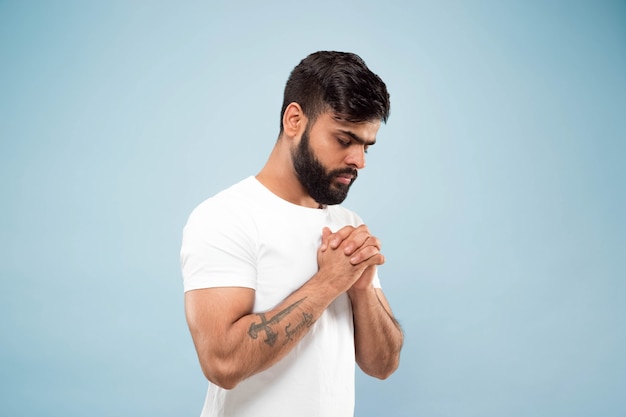 Half-length close up portrait of young hindoo man in white shirt isolated on blue background. Human emotions, facial expression, ad concept. Negative space. Standing and praying with eyes closed.
