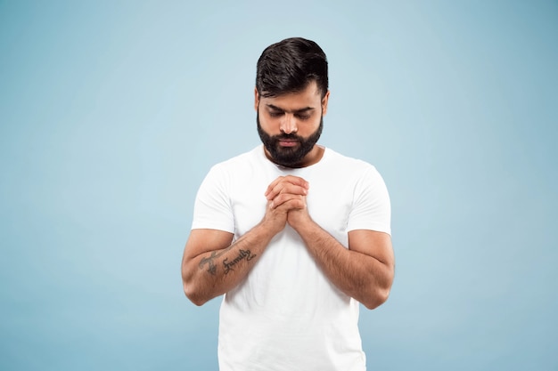Поясной конец вверх по портрету молодого человека индуизма в белой рубашке изолированной на голубой стене. Человеческие эмоции, выражение лица, концепция рекламы. Негативное пространство. Стоять и молиться с закрытыми глазами.