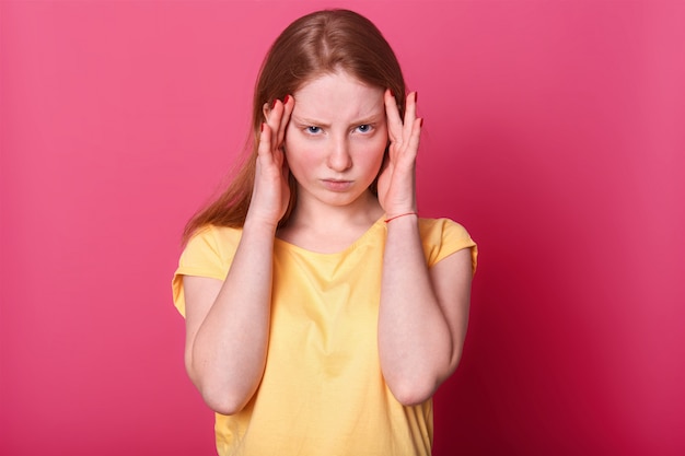 Free photo half length photo of sad teen girl with terrible headache, has serious problems in school, wears casually, posing on pink. facial expressions and people emotions concept.