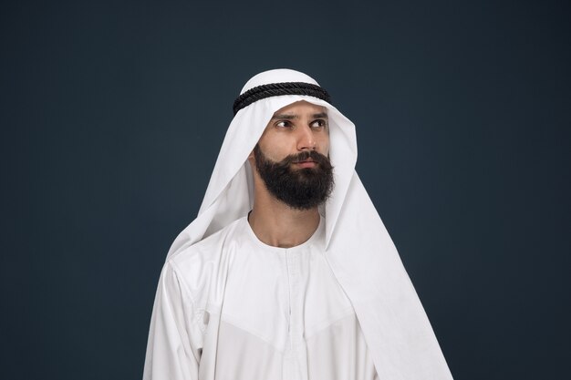 Half-length portrait of arabian saudi businessman on dark blue  space. Young male model standing and looks thoughtful