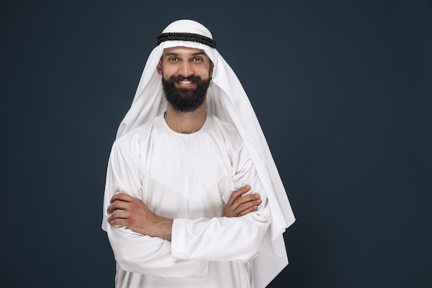 Half-length portrait of arabian saudi businessman on dark blue studio background. Young male model standing and smiling. Concept of business, finance, facial expression, human emotions.