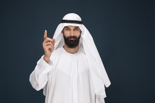 Half-length portrait of arabian saudi businessman on dark blue wall. Young male model smiling and pointing. Concept of business, finance, facial expression, human emotions.