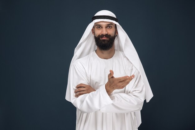진한 파란색 벽에 아라비아 사우디 사업가의 길이 초상화. 웃 고 가리키는 젊은 남성 모델. 비즈니스, 금융, 표정, 인간 감정의 개념.