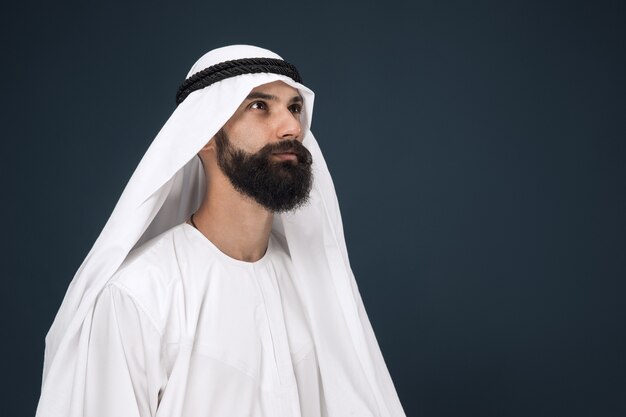 Half-length portrait of arabian saudi businessman on dark blue wall. Young male model standing and looks thoughtful. Concept of business, finance, facial expression, human emotions.