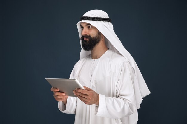 Half-length portrait of arabian saudi businessman on dark blue wall. Young male model using tablet or gadget. Concept of business, finance, facial expression, human emotions, technologies.