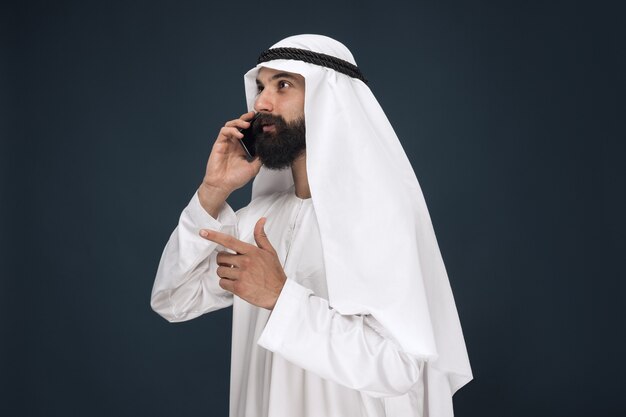 Поясной портрет арабского саудовца на темно-синей стене студии. Мужская модель с помощью смартфона, звонит. Понятие бизнеса, финансов, мимики, человеческих эмоций, технологий.