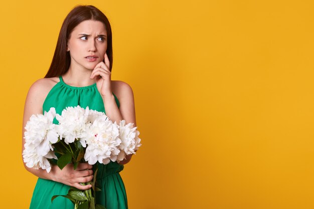 Half length portrait of attractive brunette woman in green sundress holding bouquet of white peonies on yellow, looking pensivly aside, keeps finger on cheek, copy space for advertisment.