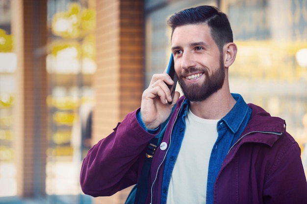 Foto gratuita ritratto a mezza lunghezza di un bell'uomo hipster con barba, maglietta bianca, camicia blu e giacca marrone che tiene in mano uno smartphone.