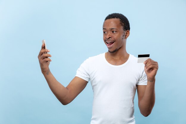 Half-length portrait of young african-american man in white shirt holding a card and smartphone on blue space