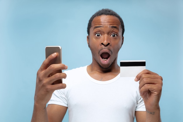 Half-length portrait of young african-american man in white shirt holding a card and smartphone on blue space