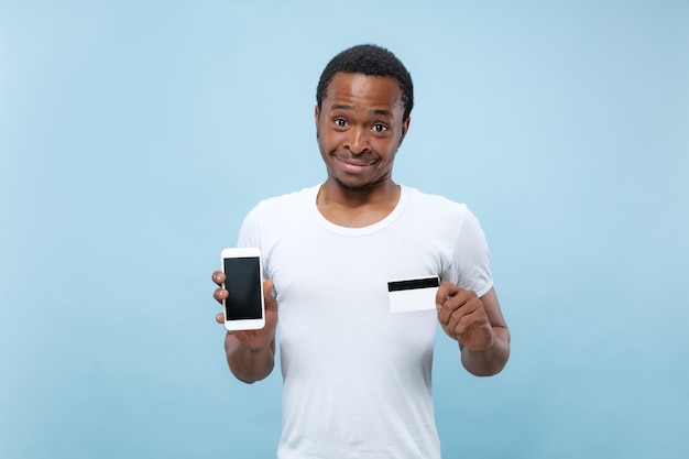 Half-length portrait of young african-american man in white shirt holding a card and smartphone on blue space