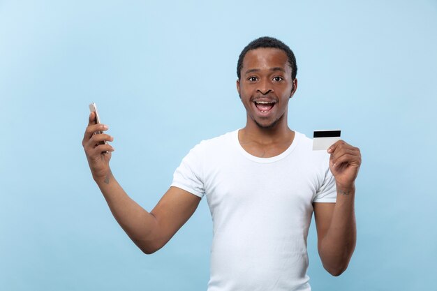 Half-length portrait of young african-american man in white shirt holding a card and smartphone on blue space