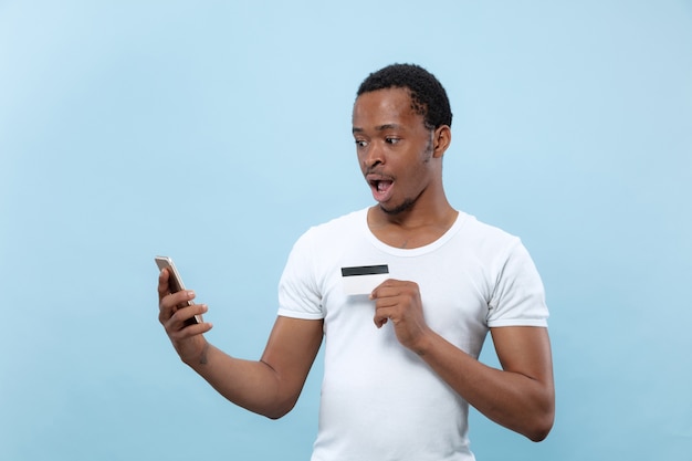 Half-length portrait of young african-american man in white shirt holding a card and smartphone on blue wall. Human emotions, facial expression, ad, sales, finance, online payments concept.