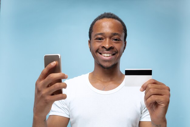 Half-length portrait of young african-american man in white shirt holding a card and smartphone on blue wall. Human emotions, facial expression, ad, sales, finance, online payments concept.