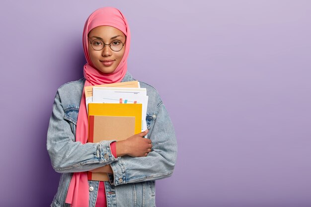 魅力的な自信を持っているイスラム教徒の大学生の半分の長さのショットは、ノート、紙の文書を保持し、レッスンのプロジェクト作業を準備し、ピンクのヒジャーブ、丸い眼鏡、ジーンズの服を着ています。概念を学ぶ