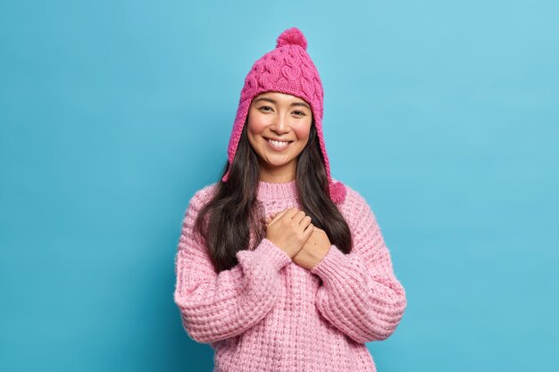 Half length shot of beautiful Asian woman in knitwear being thankful for heartwarming words smiles pleasantly poses against blue wall