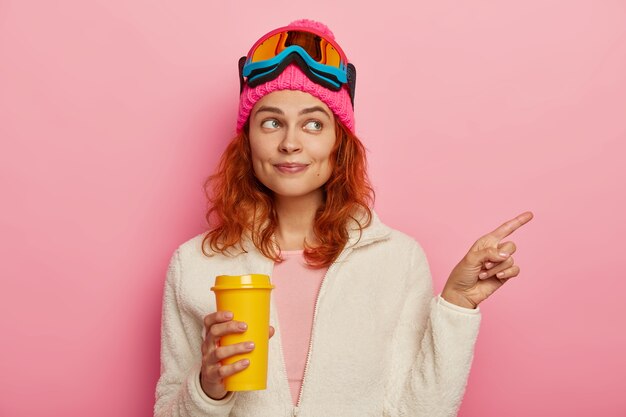 Half length shot of beautiful redhead girl points right, shows direction to tourist in resort place, has active winter rest, drinks takeaway coffee, wears ski mask, isolated over pink background