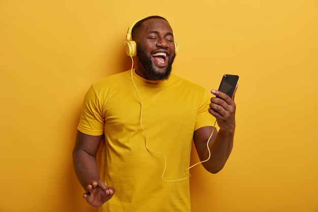 Half length shot of black guy listens music to chill, holds modern smartphone and wears headphones on ears, enjoys nice track, poses against yellow background.