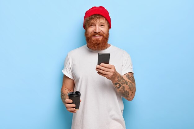 쾌활한 수염 난 빨간 머리 남자의 절반 길이 샷은 세련된 모자, 흰색 캐주얼 티셔츠를 입고 휴대 전화, 테이크 아웃 커피를 들고 좋은 분위기에 있으며 메시지를 입력합니다.