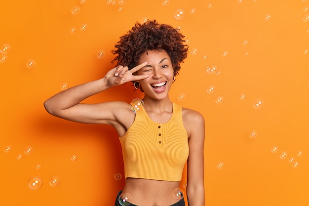 Half length shot of curly haired Afro American woman in good physical shape wears cropped top makes peace gesture over eye smiles broadly isolated over orange wall
