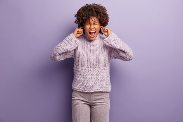 Half length shot of depressed mixed race woman plugs ears, doesnt want to hear quarrel, annoyed of gossips, wears knitted jumper and trousers, stands over purple  wall. Ignoring noise