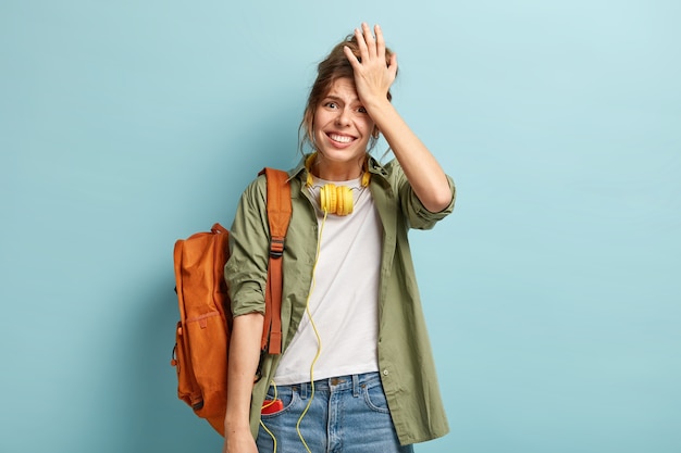 Half length shot of displeased woman keeps hand on forehead, regrets something, looks in dissatisfaction, has headphones on neck