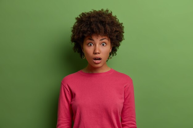 Половинный снимок смущенной безмолвной женщины с афро-волосами, у которой глаза прикованы к страху, удивленно открывает рот, реагирует на поразительные новости, одетая в розовый джемпер, изолирована на зеленой стене