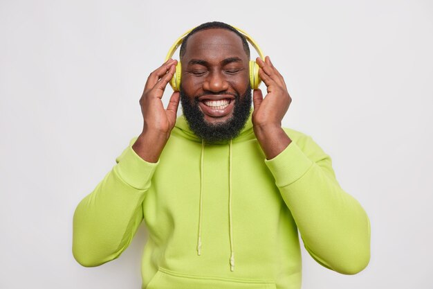 Half length shot of handsome cheerful bearded guy with dark skin listens to music in wireless headphones smiles toothily has perfect white teeth wears green sweatshirt isolated over white wall