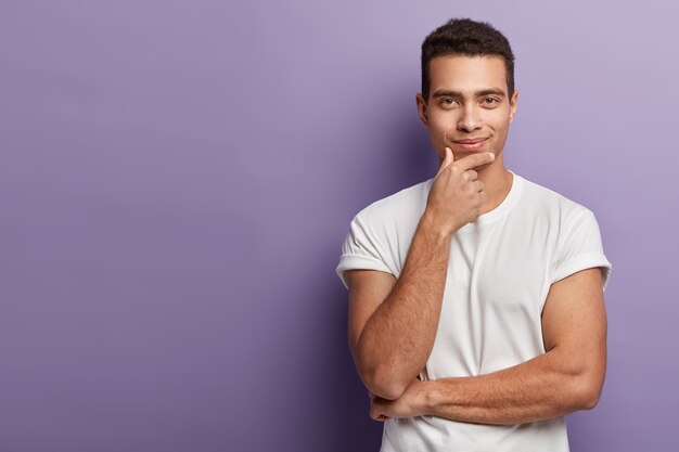 Снимок в половину длины: красивый европейский мужчина держит подбородок, смотрит прямо с довольным уверенным выражением лица, в белой футболке, позирует над фиолетовой стеной, свободное пространство оставлено в стороне
