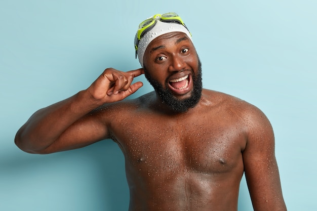 Half length shot of happy African American man has water in ear after diving, wet dark skin, opens mouth widely