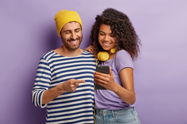 Half length shot of happy girlfriend and boyfriend watch funny video content on smartphone, stand closely, have cheerful expressions, connected to wireless internet