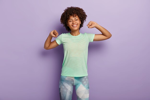 Half length shot of happy young Afro American woman stretches hands, does morning exercises with good mood