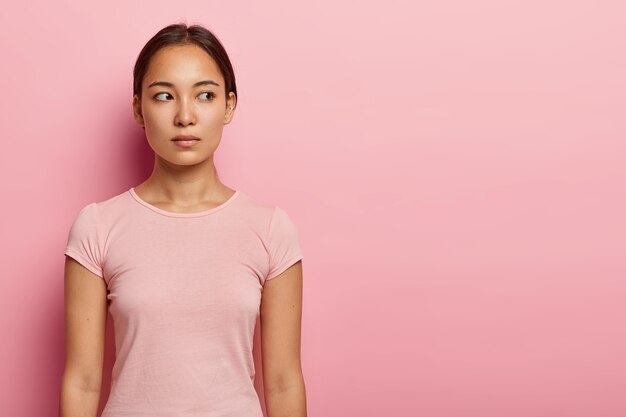 사랑스러운 진지한 여성의 절반 길이 샷은 빈 공간에 옆으로 보이고 잠겨있는 표정, 건강한 피부 및 특정 외모를 가지고 있으며 분홍색 벽에 고립 된 캐주얼 티셔츠를 입습니다. 민족성 개념