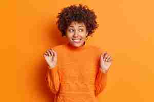 無料写真 ポジティブなアフリカ系アメリカ人女性が唇を噛み、手を上げたままの半分の長さのショットが不思議なことに離れて見える笑顔興味をそそられる何か面白いものがオレンジ色の壁に隔離されたカジュアルなジャンパーを着ている