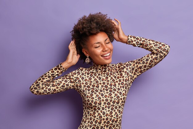 Half length shot of optimistic curly haired woman moves carefree, smiles pleasantly, has eyes closed, touches head, imagines something very pleasant