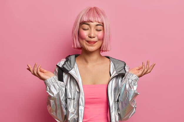 Half length shot of pleased Asian woman raises hands, stands with eyes closed, feels happy, imagines something pleasant, wears silver jacket 