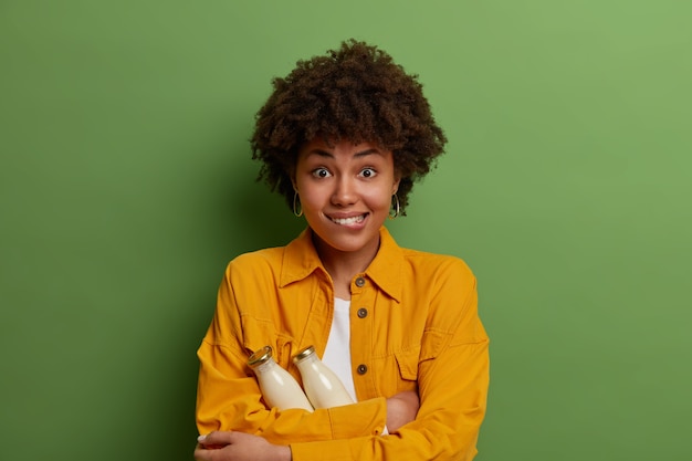 Half length shot of positive Afro American woman bites lips, holds two glass bottles of plant based milk, drinks only organic beverage, keeps to healthy diet, wears yellow shirt, stands indoor