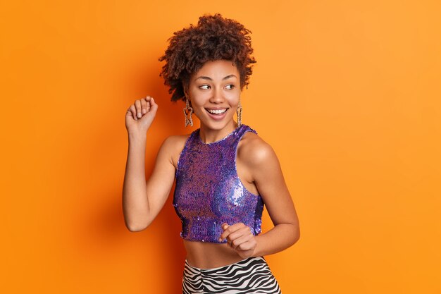 Half length shot of positive carefree curly woman dances and moves shakes arms wears cropped top earrings smiles broadly isolated over vivid orange wall