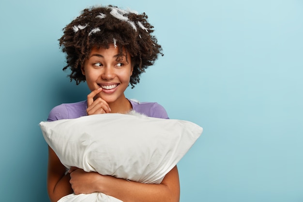 Free photo half length shot of positive dark skinned girl looks with dreamy expression, keeps hand near mouth, smiles sincerely, has pleasant dream at night, holds pillow, prepares for sleep, isolated on blue