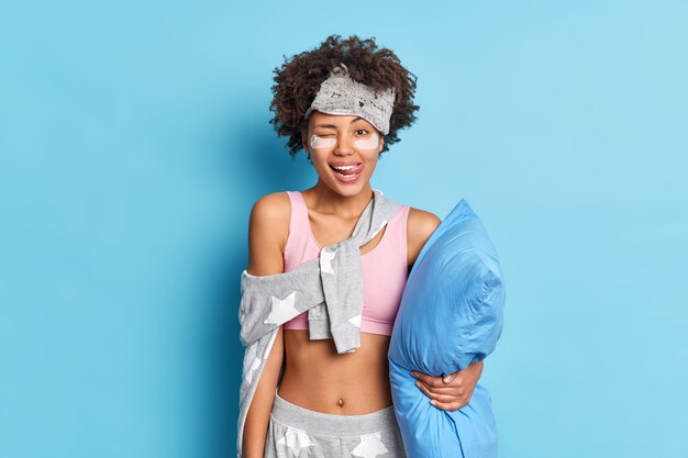 Half length shot of positive girl shows tongue winks eye has happy mood going to sleep holds pillow dressed in pajama undergoes beauty treatments at home isolated on blue studio wall