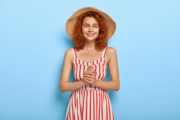 生姜髪型の可愛らしい笑顔の女性のハーフレングスショット、麦わら帽子とストライプのドレスを着て、彼氏とのデートの準備ができて、手をつないで、満足のいく表情をしています。夏のファッション