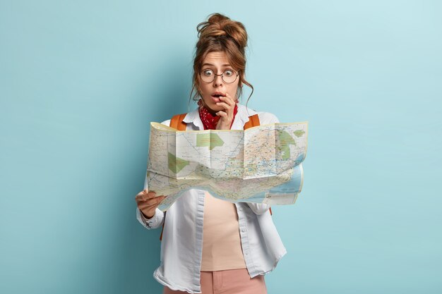 Half length shot of scared embarrassed woman looks stressfully at map, afraids of being lost, wears optical glasses