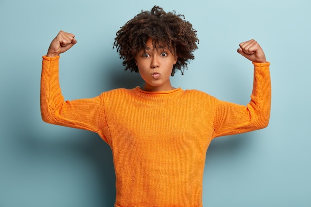 Half length shot of serious Afro American woman with curly hair, raises hands, shows muscles, tries to be strong